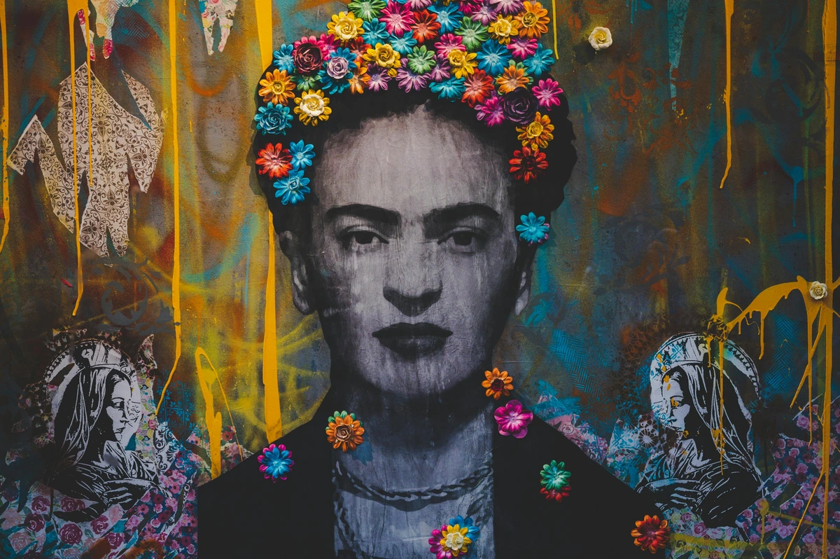 Vibrant graffiti mural featuring a portrait of Frida Kahlo adorned with a colorful floral crown, set against a textured background with yellow paint drips.