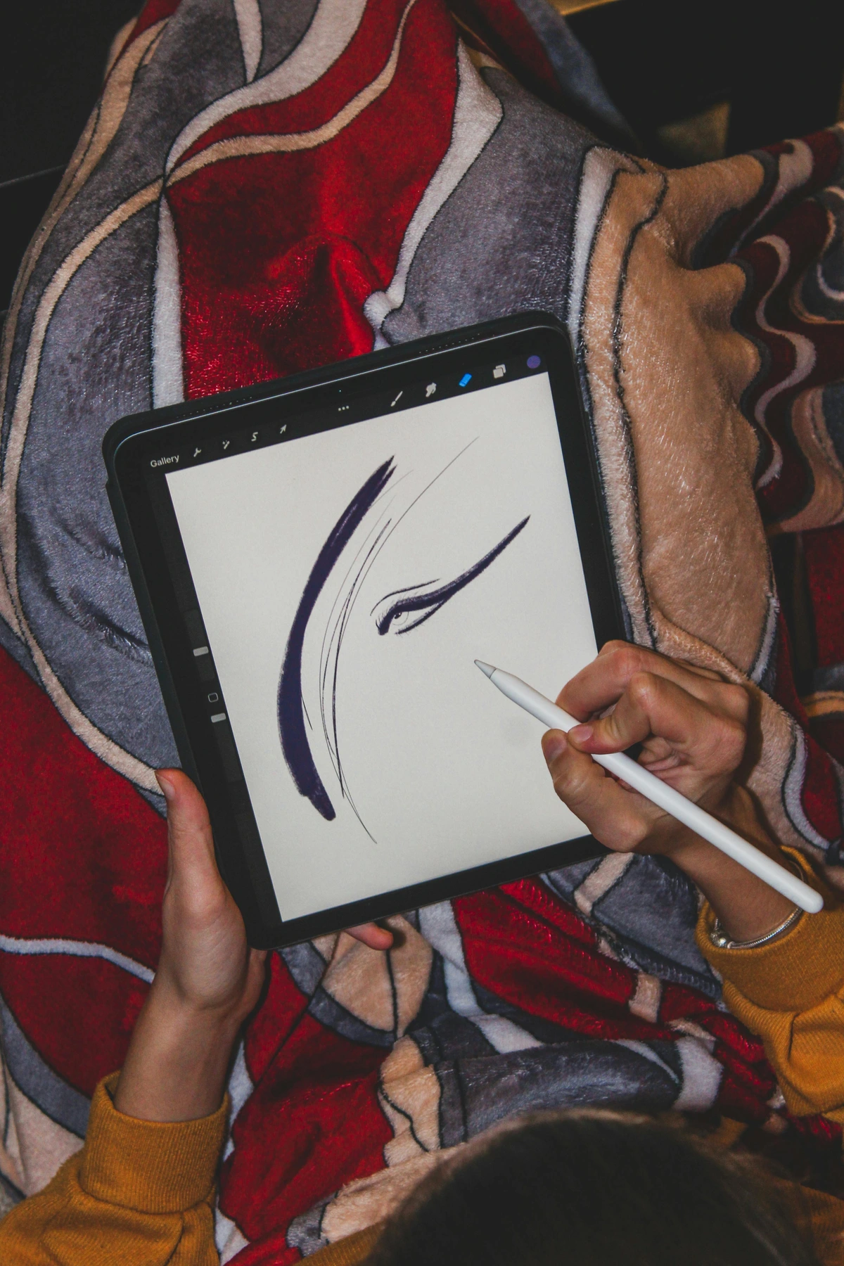 A person's hands holding a stylus and drawing an abstract eye sketch on a digital tablet, covered by a patterned blanket.