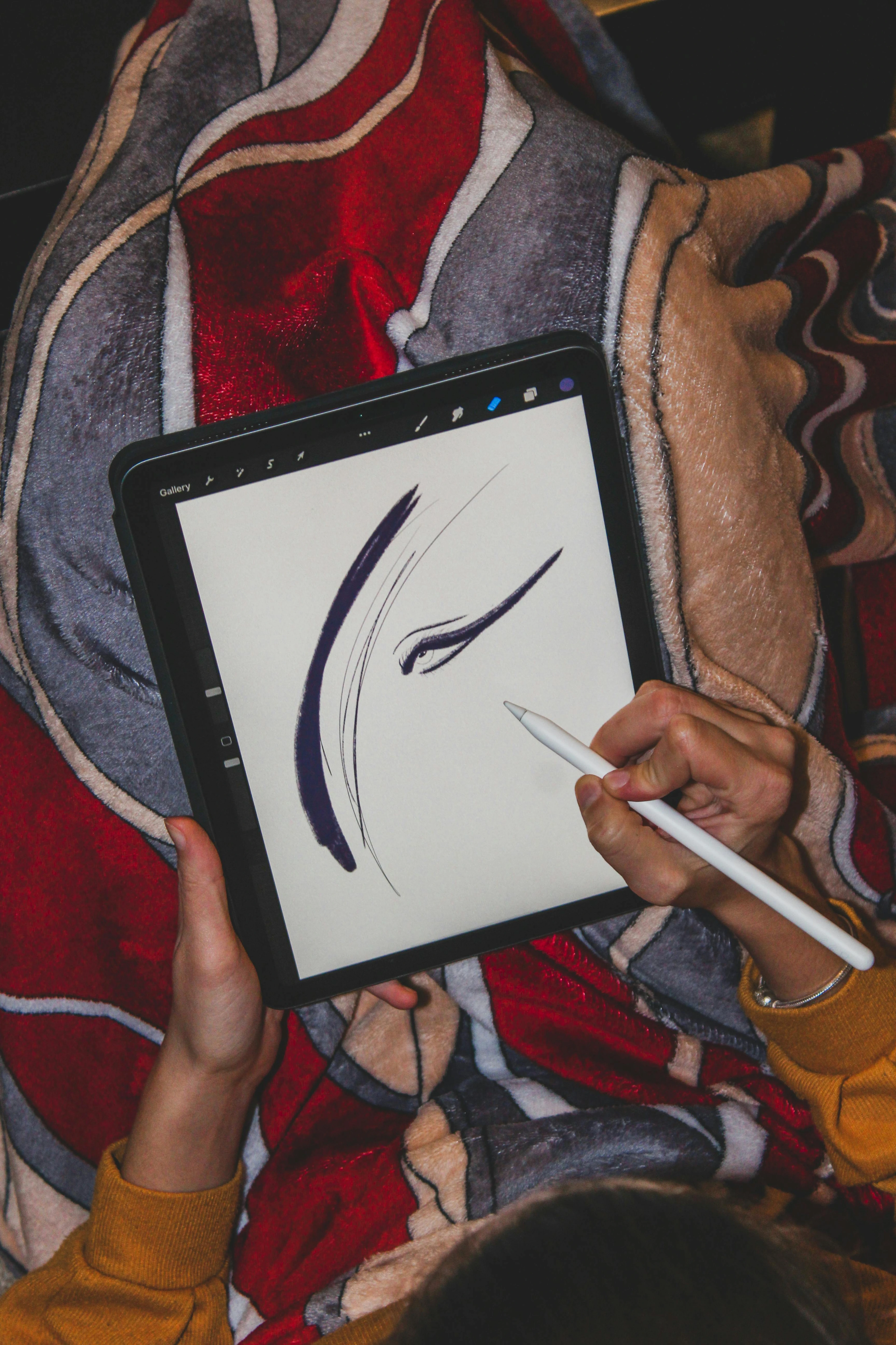 A person's hands holding a stylus and drawing an abstract eye sketch on a digital tablet, covered by a patterned blanket.