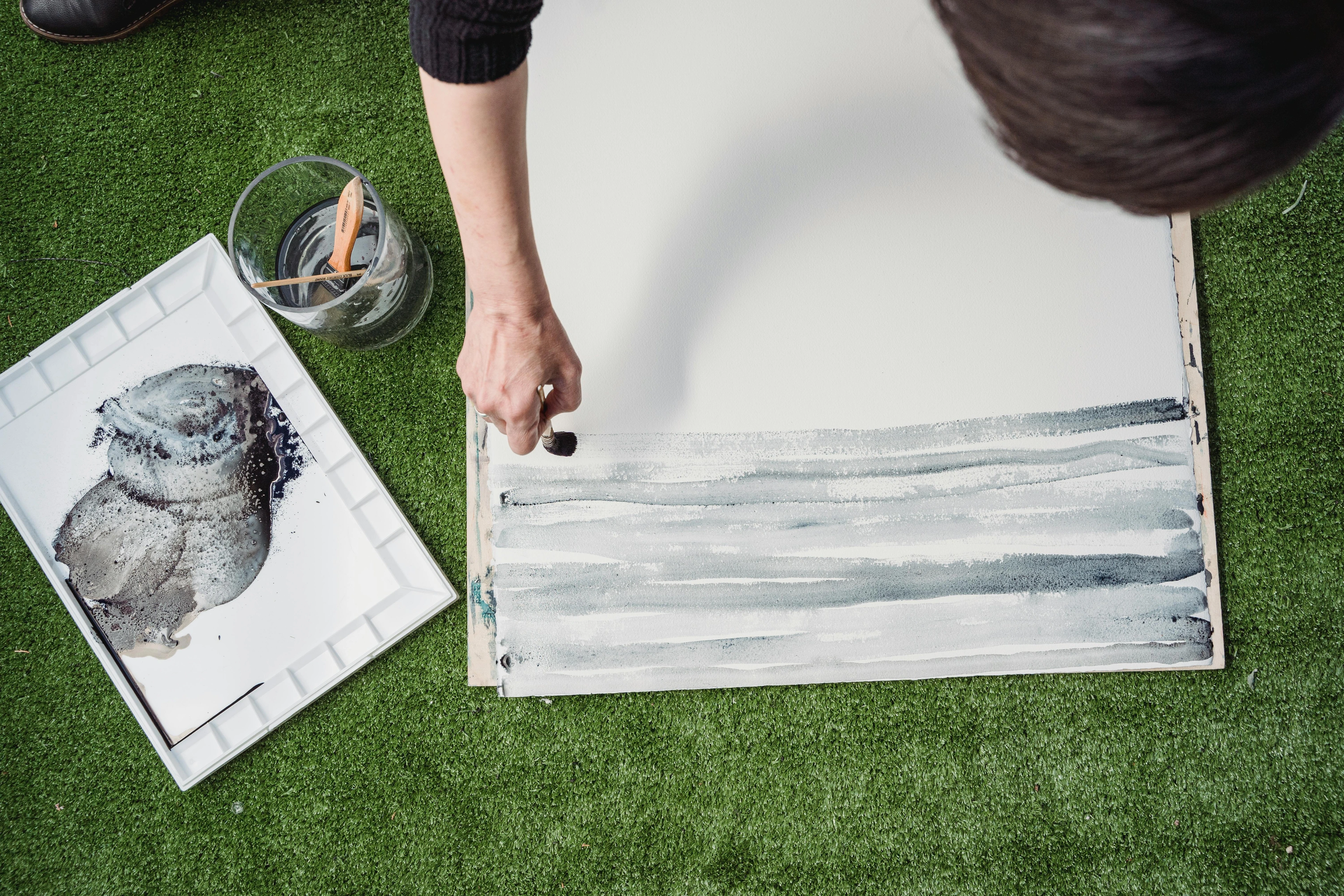 High-angle shot of a person applying grey watercolor washes to a canvas, creating abstract horizontal lines. A palette with paint and brushes is visible on the side.
