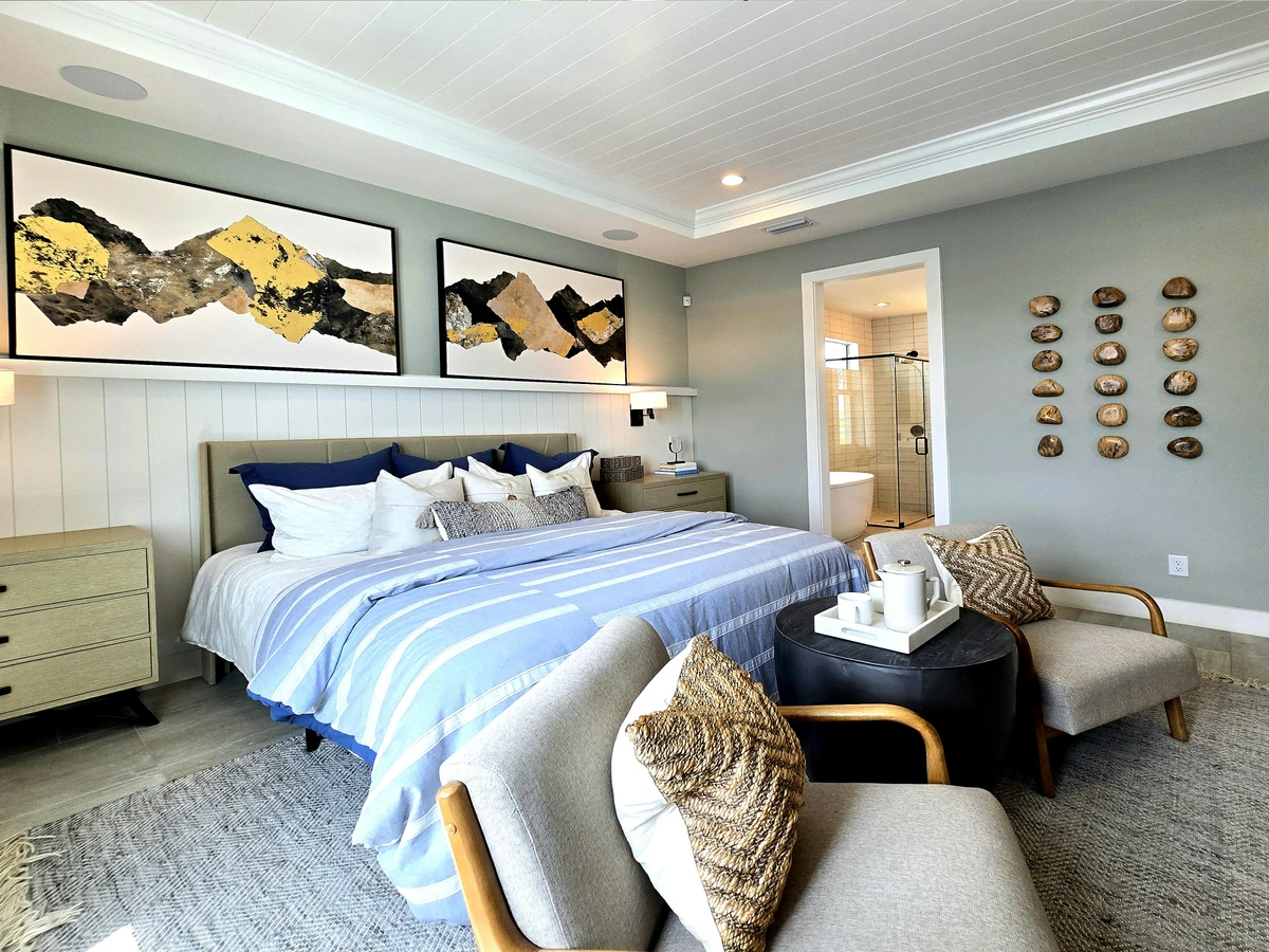 Modern bedroom featuring abstract mountain landscape art above a bed with blue and white striped bedding, flanked by nightstands and accent chairs.