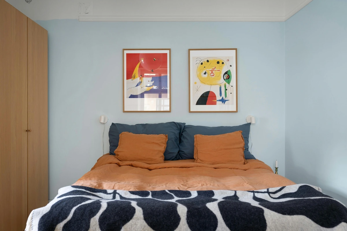 Two framed abstract art pieces hanging on a light blue wall above a bed with orange and blue bedding and a black and white patterned blanket.