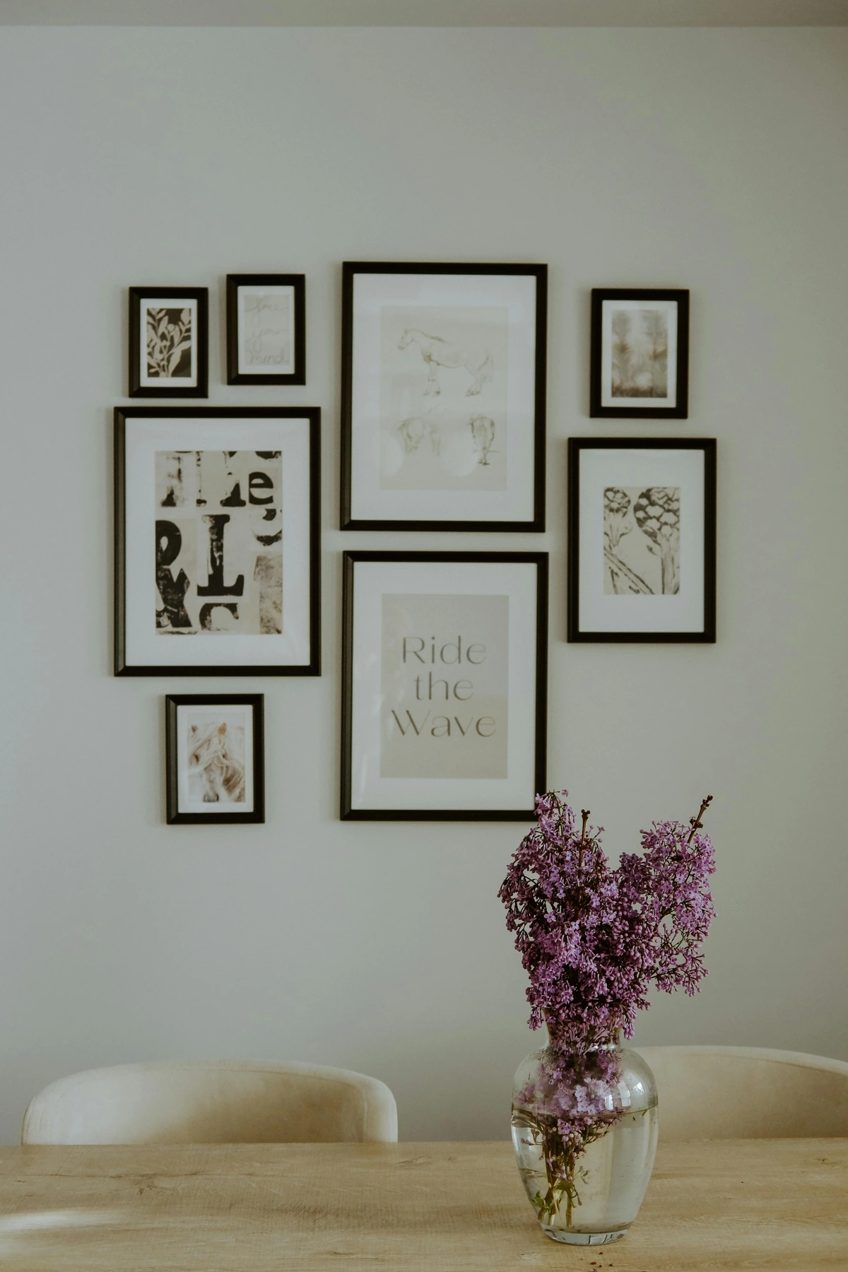 A gallery wall of framed art above a wooden dining table with a vase of purple lilac flowers.