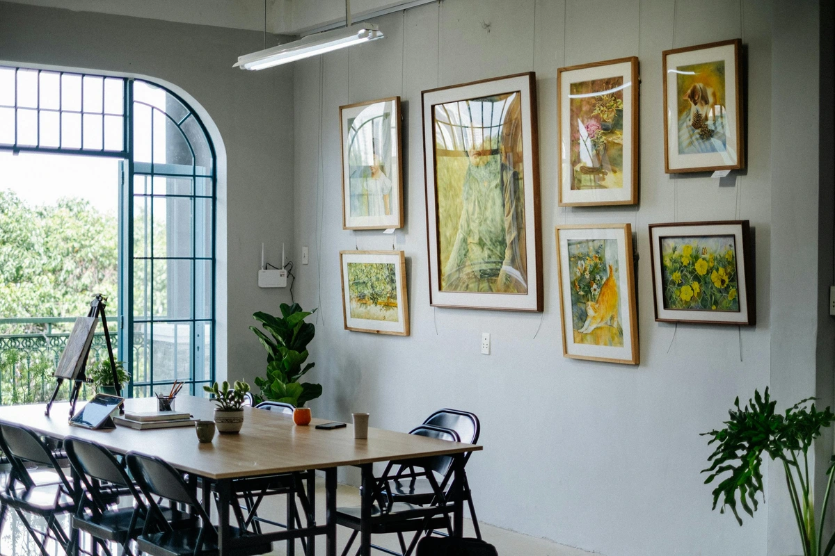 Modern dining room with a large wooden table, black chairs, and a gallery wall of paintings, featuring an arch window with natural light and indoor plants.