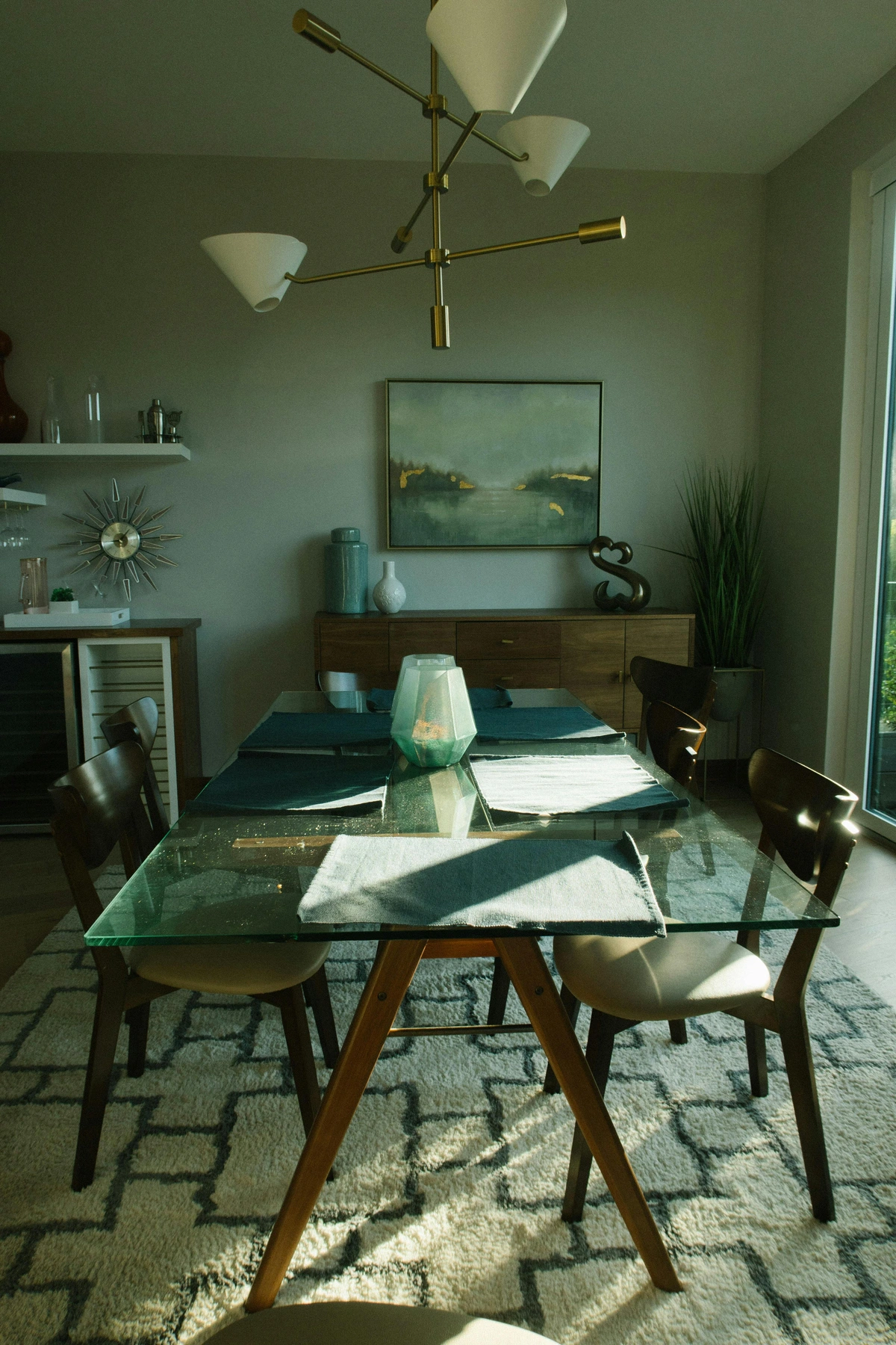 A modern dining room with a glass-top table, wooden chairs, and abstract wall art, illuminated by natural sunlight.