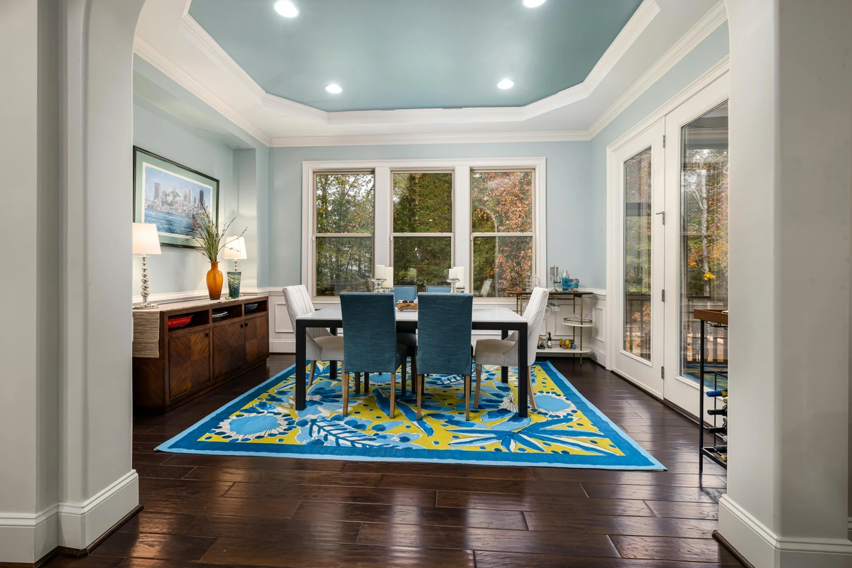 A modern dining area with a blue and yellow floral rug, blue upholstered chairs, and a white marble-top dining table, illuminated by natural light from large windows.