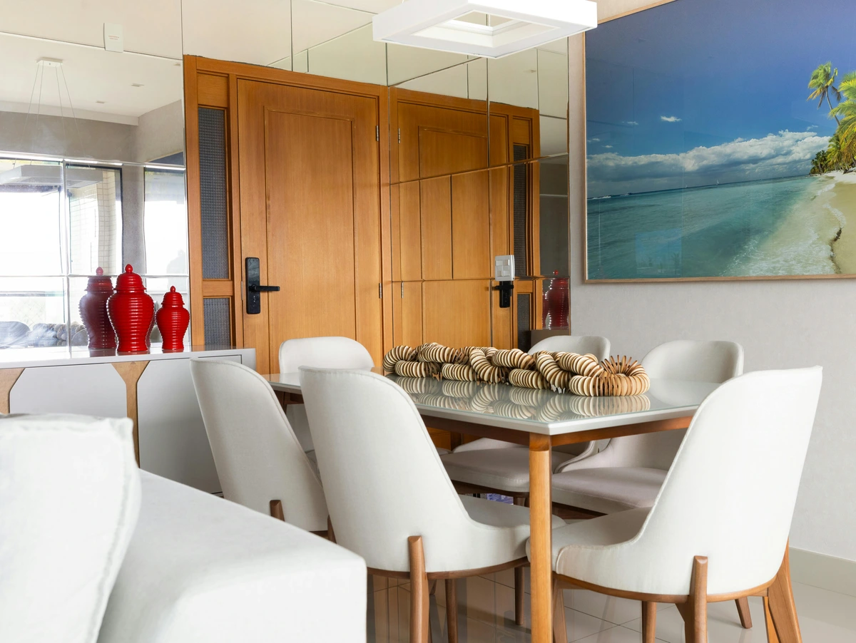 Modern dining room featuring a glass-top dining table with white chairs, adorned with a tropical beach artwork and red vases on a sideboard.