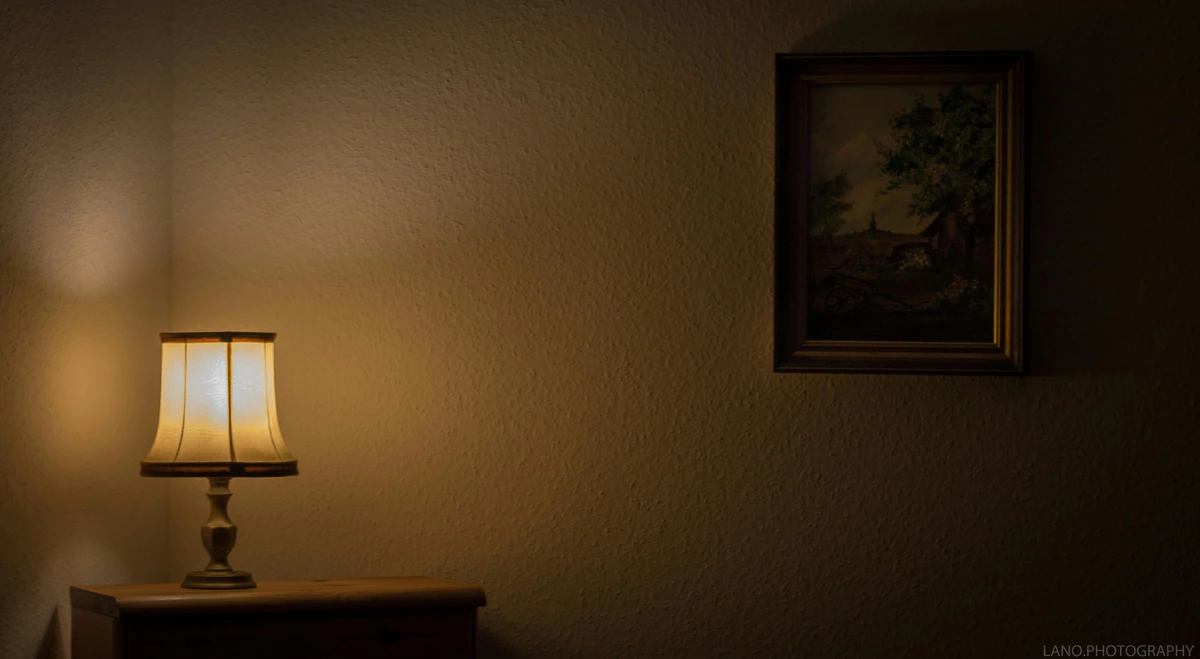 A warmly lit table lamp on a wooden nightstand in a dimly lit room, with a framed landscape painting on the textured wall.