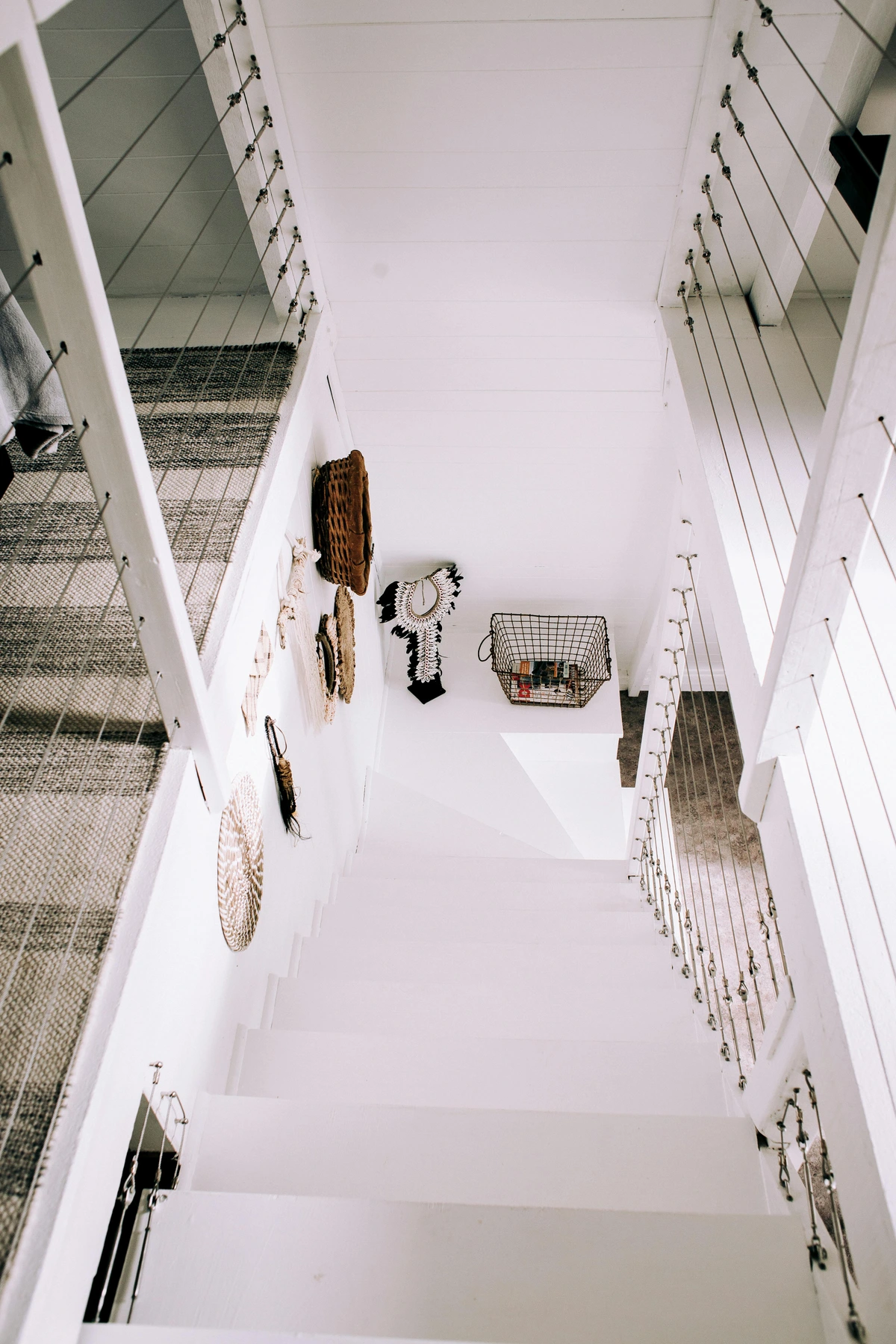 Modern minimalistic staircase with woven wall art and a wire basket, featuring white walls and cable railings.