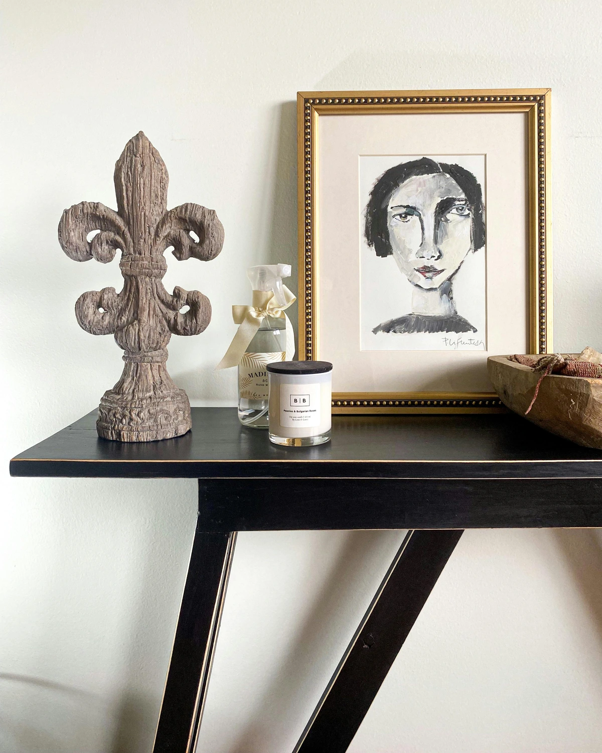 Stylish console table with a framed portrait painting, a fleur de lis sculpture, a scented candle, and a decorative spray bottle.