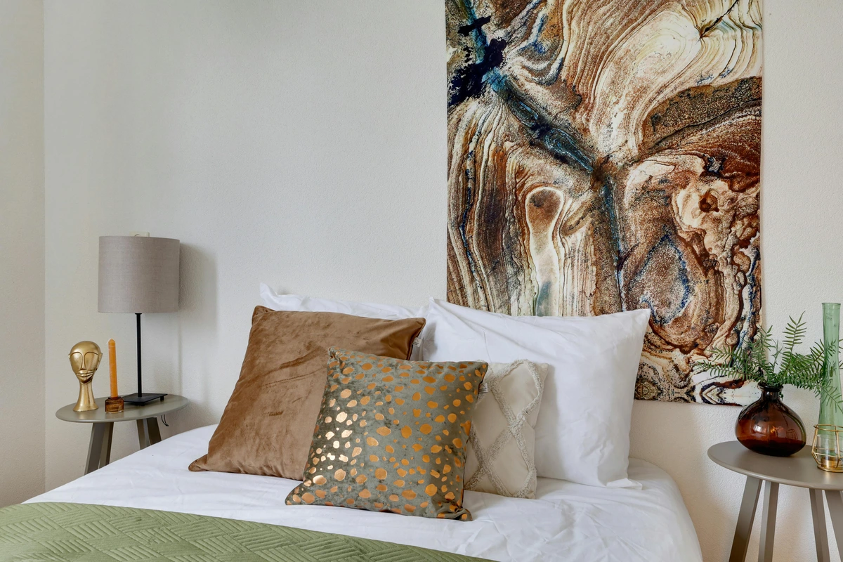 Bedroom with a large abstract painting above the bed, featuring warm earth tones and blue accents. The bed is made with white linens and several decorative pillows, including a brown velvet pillow and a grey pillow with gold spots. A green quilt is folded at the foot of the bed. Beside the bed are nightstands with a lamp, a candle, and a vase with greenery.