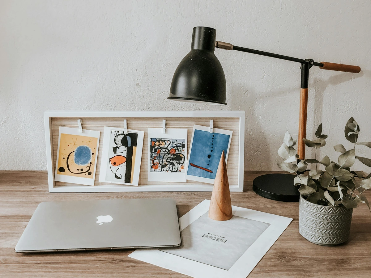 Modern home office desk setup with a laptop, abstract art prints in a white frame, a black desk lamp, and a potted eucalyptus plant.
