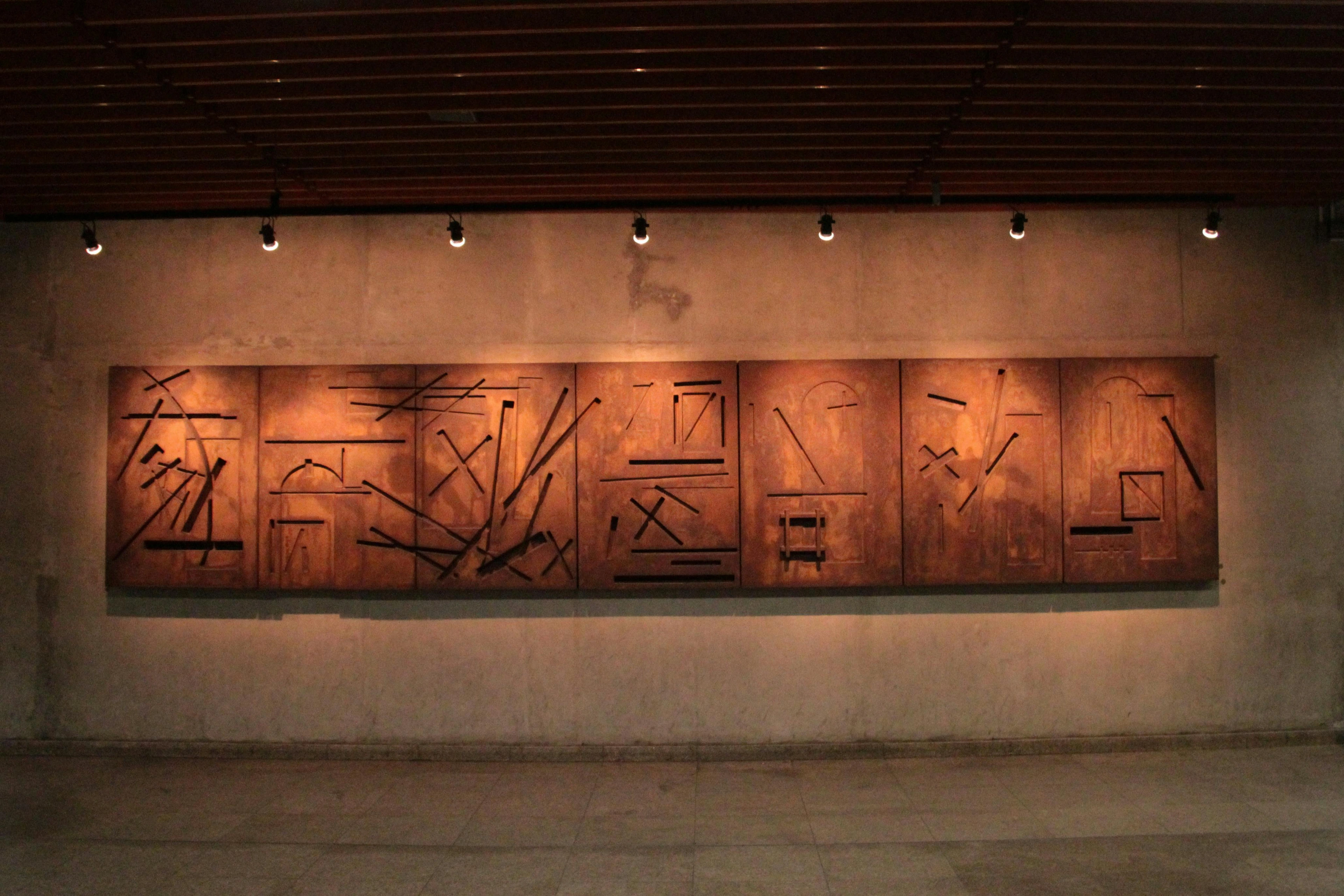 Abstract wooden wall art with geometric shapes and lines, mounted on a textured wall with spotlights.