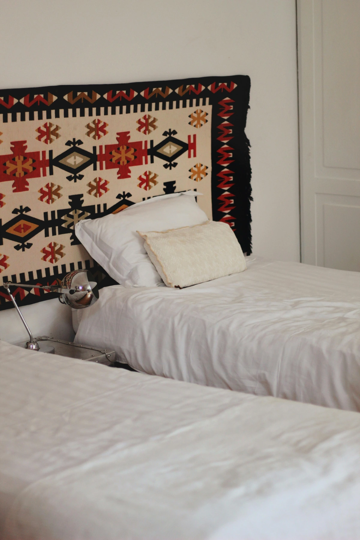 Hotel room interior featuring two twin beds with white bedding, a decorative tapestry above the headboards, and a chrome table lamp.
