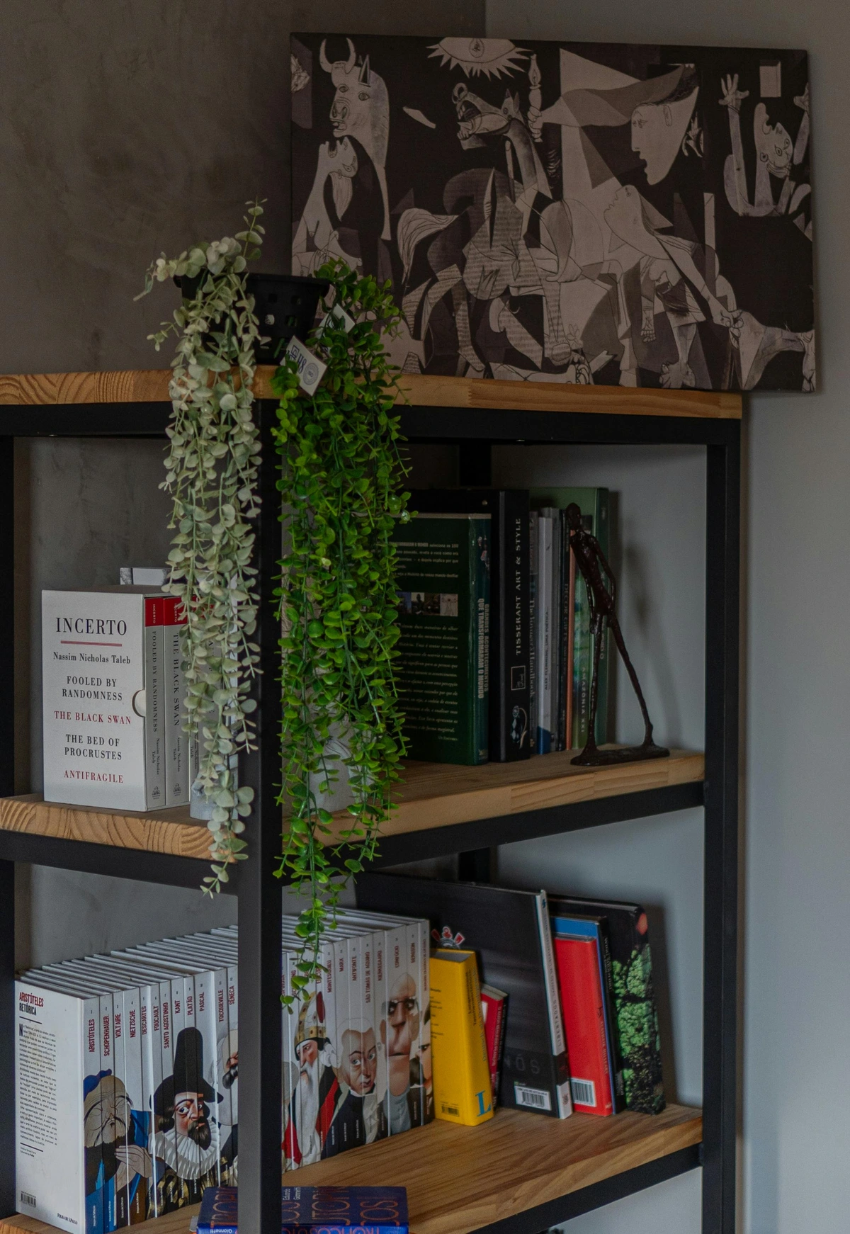 A framed print of Picasso's Guernica painting hangs above a wooden bookshelf filled with books and artificial plants.
