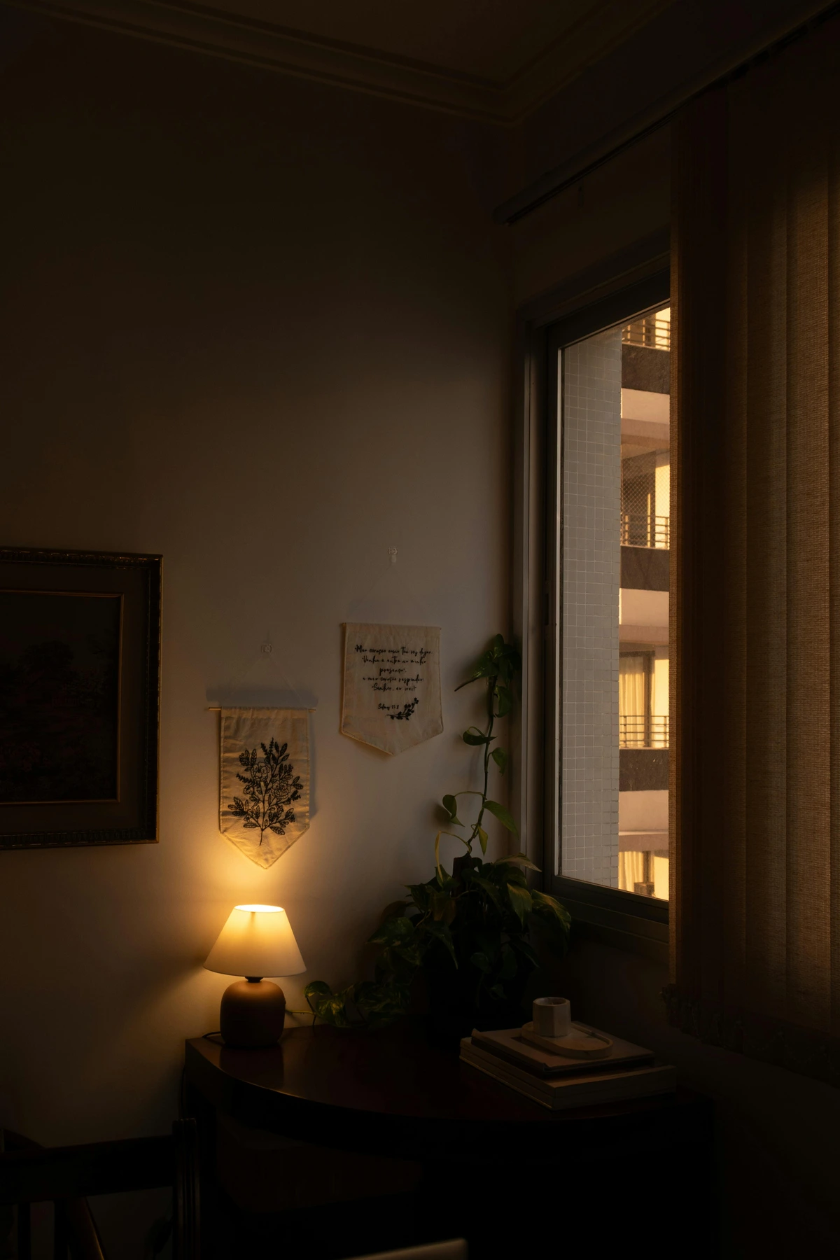 Warm ambient light from a table lamp illuminates a cozy indoor corner with books, a plant, and wall art, with a view of a building through the window.