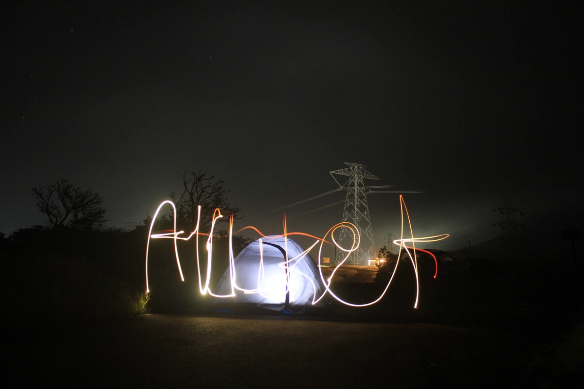 Light painting of the word 'Allure' in front of a glowing tent at night, with power lines and a pylon in the background.