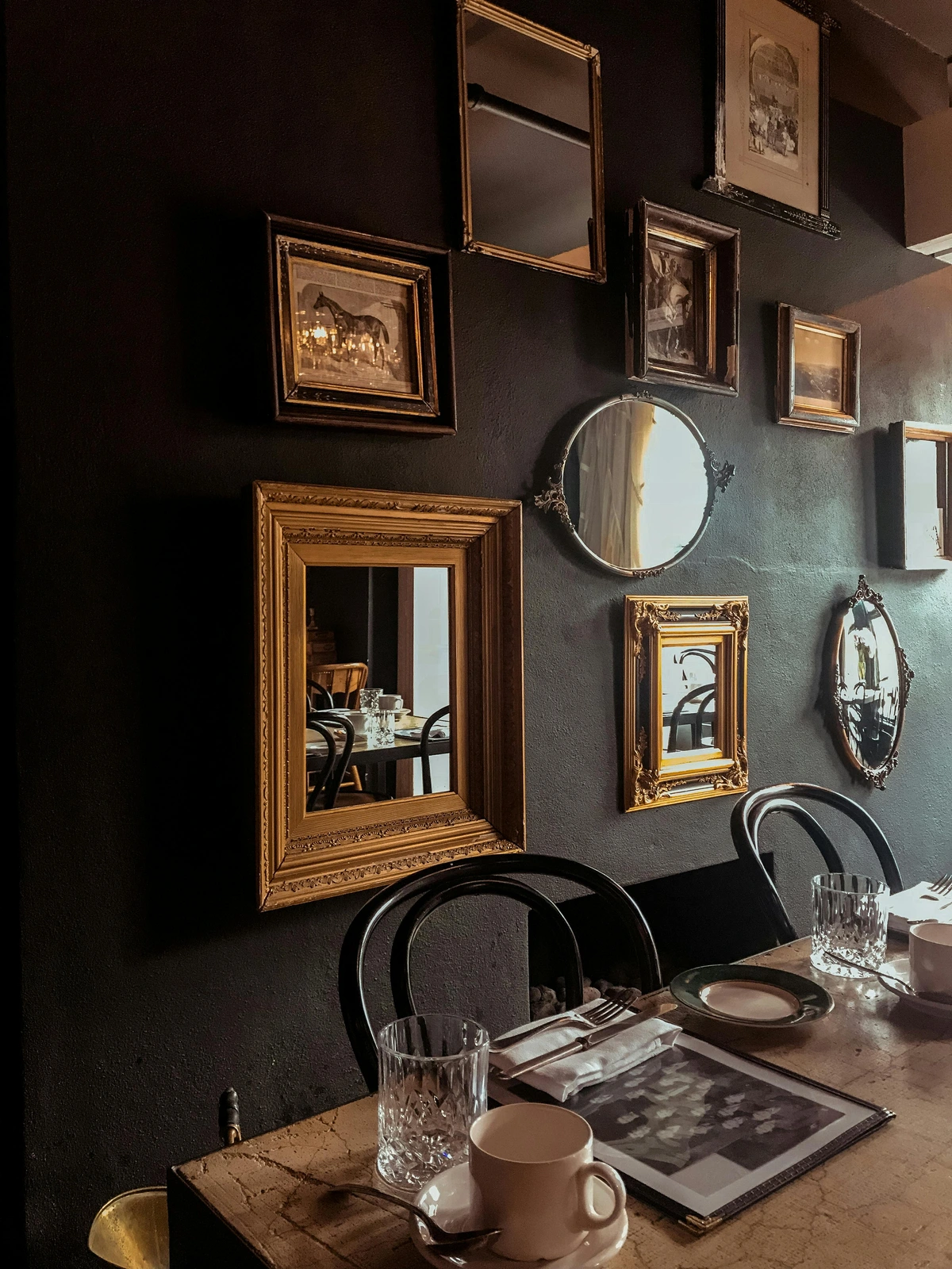 Collection of framed mirrors and art pieces arranged on a dark, textured wall above a dining table.