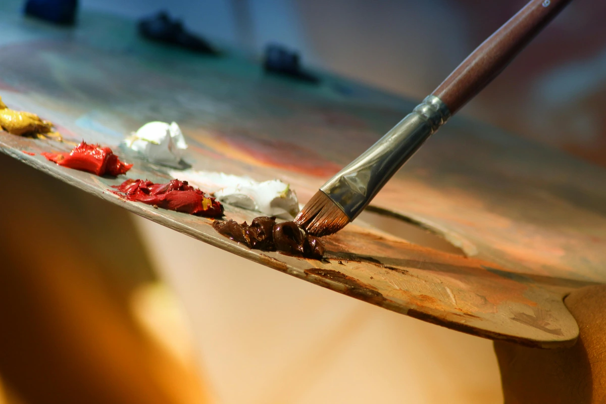 Close-up of a paintbrush picking up dark brown paint from an artist's palette, with other colors like red and white visible, illustrating the tools and craft behind still life creation.