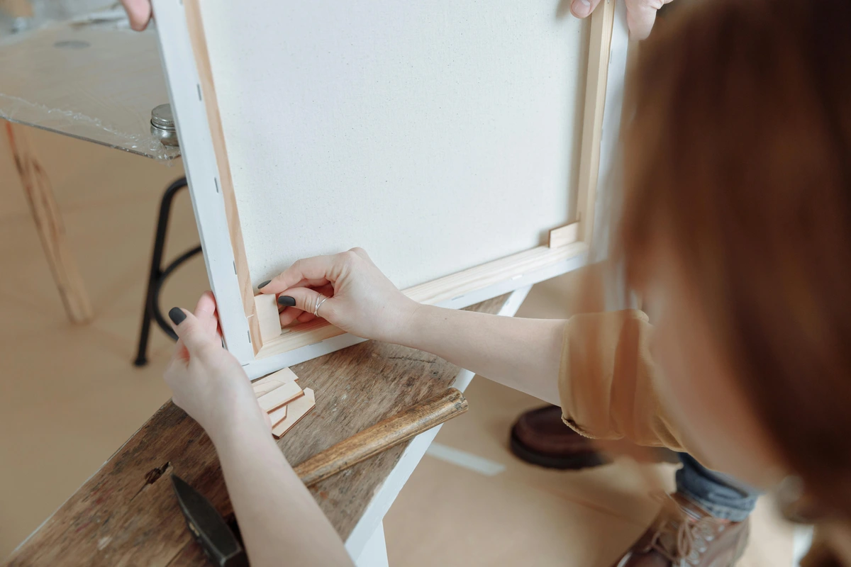 A woman's hands carefully stretching a blank canvas onto wooden stretcher bars, preparing it for painting.