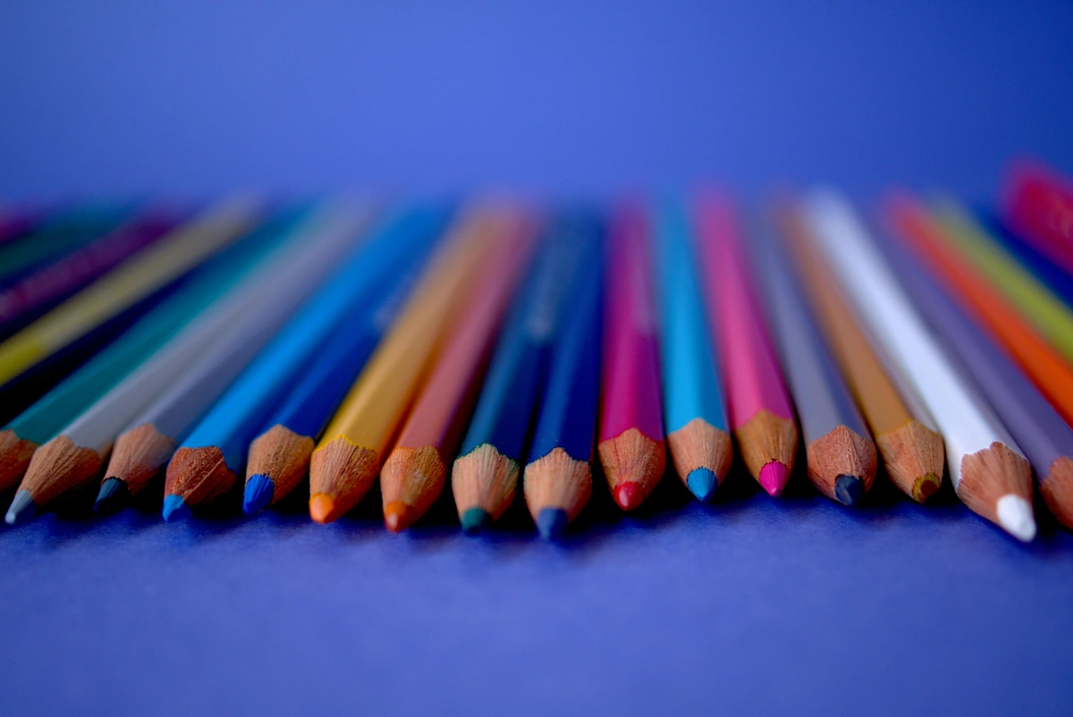 Assorted color colored pencils arranged in a row on a blue background, sharp tips in focus.