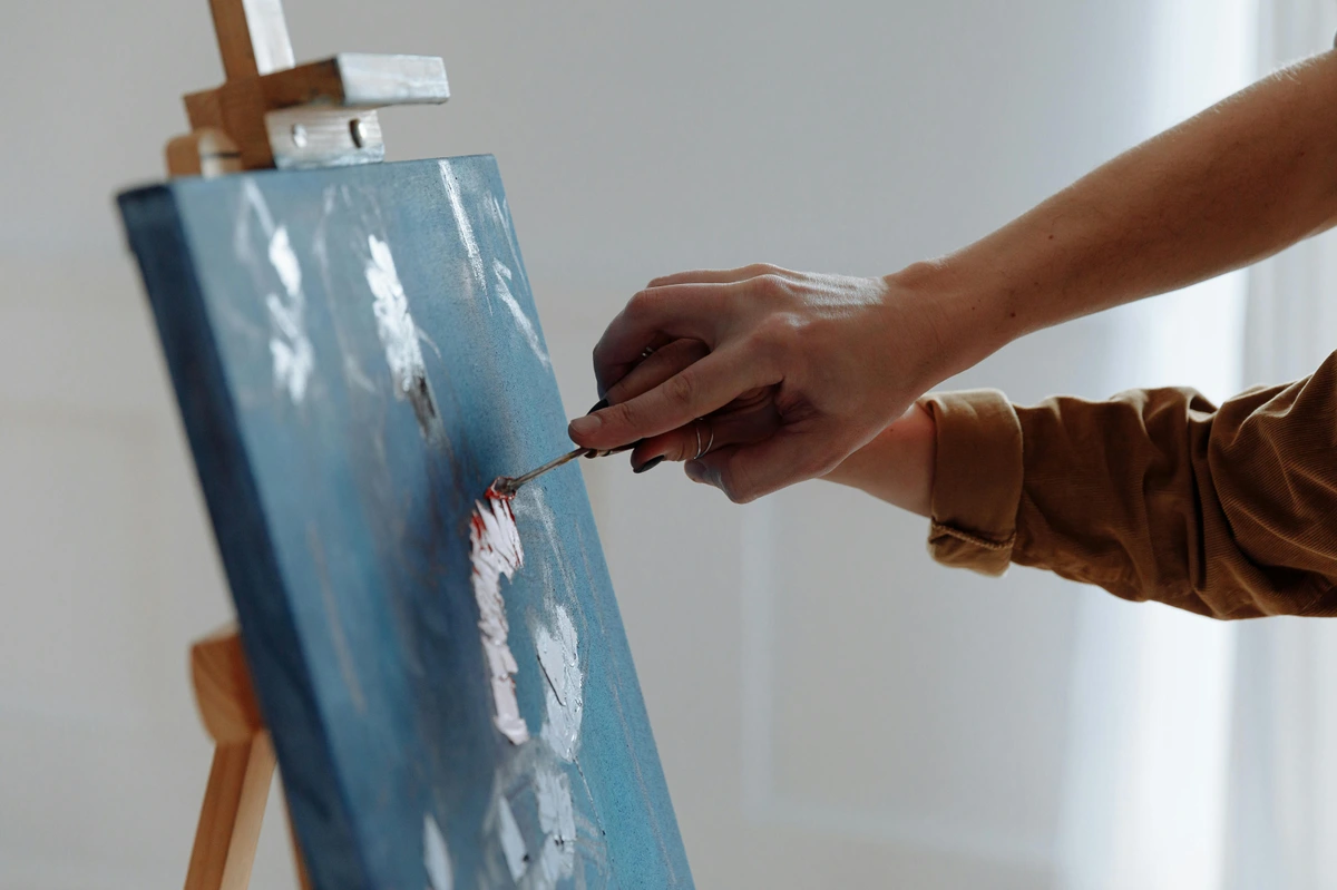 Artist painting close-up: hands using a palette knife on a blue canvas with white paint strokes