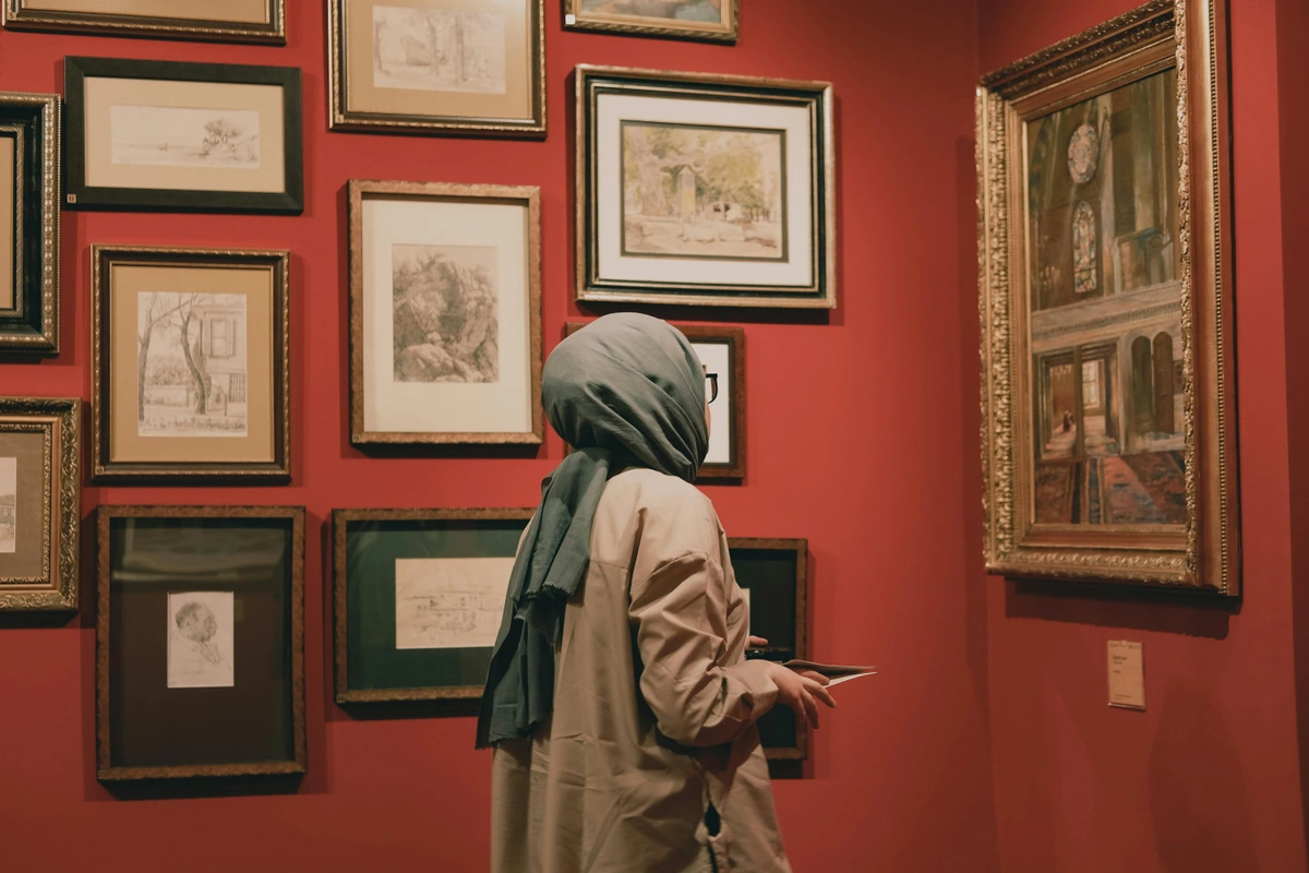 Woman wearing a hijab and a beige coat looking at paintings displayed on a red wall in an art museum.
