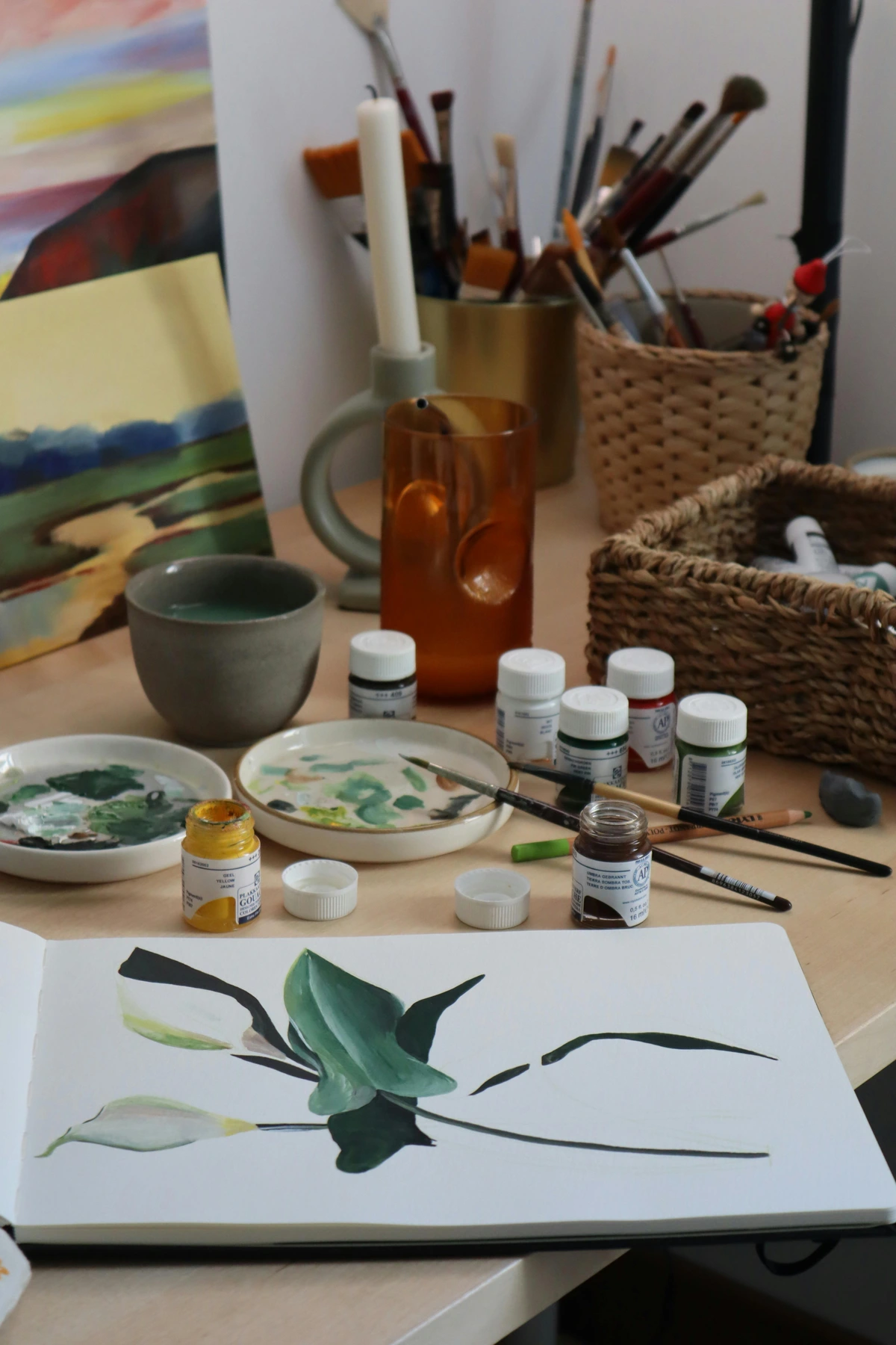 Gouache paint bottles, brushes, and a painting in progress on a wooden table