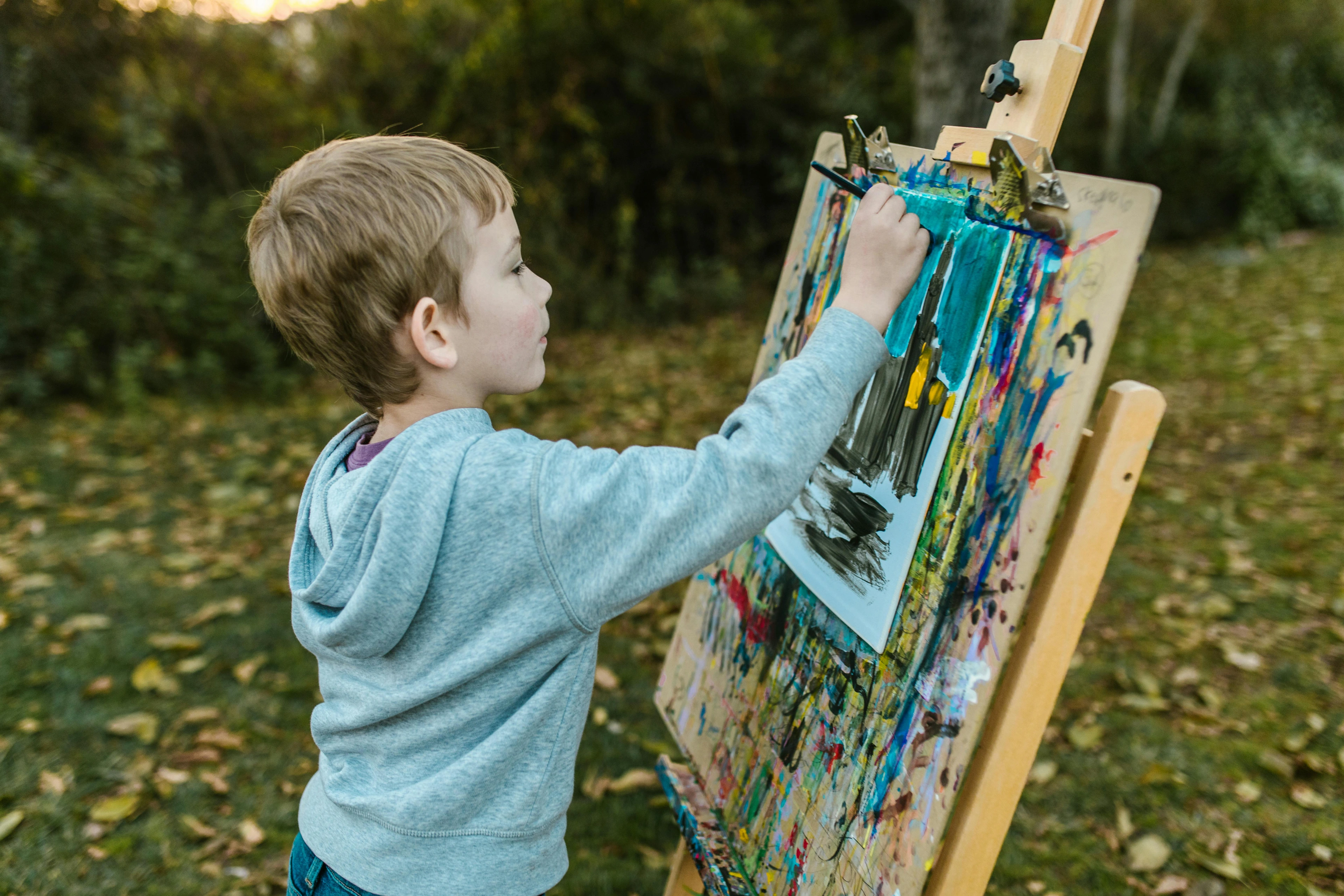 A young boy is focused on painting a colorful picture on an easel outdoors, with autumn leaves on the ground.