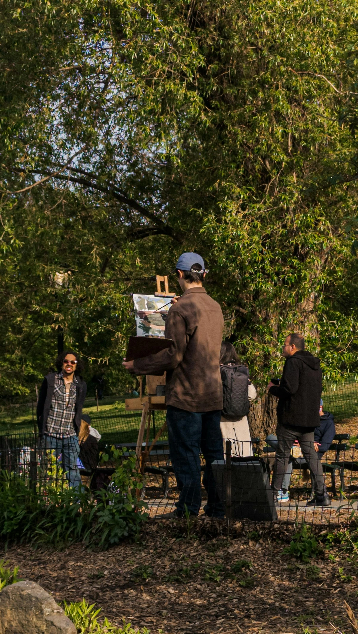 Artist painting outdoors in a park with a portable easel, surrounded by trees and other people observing.
