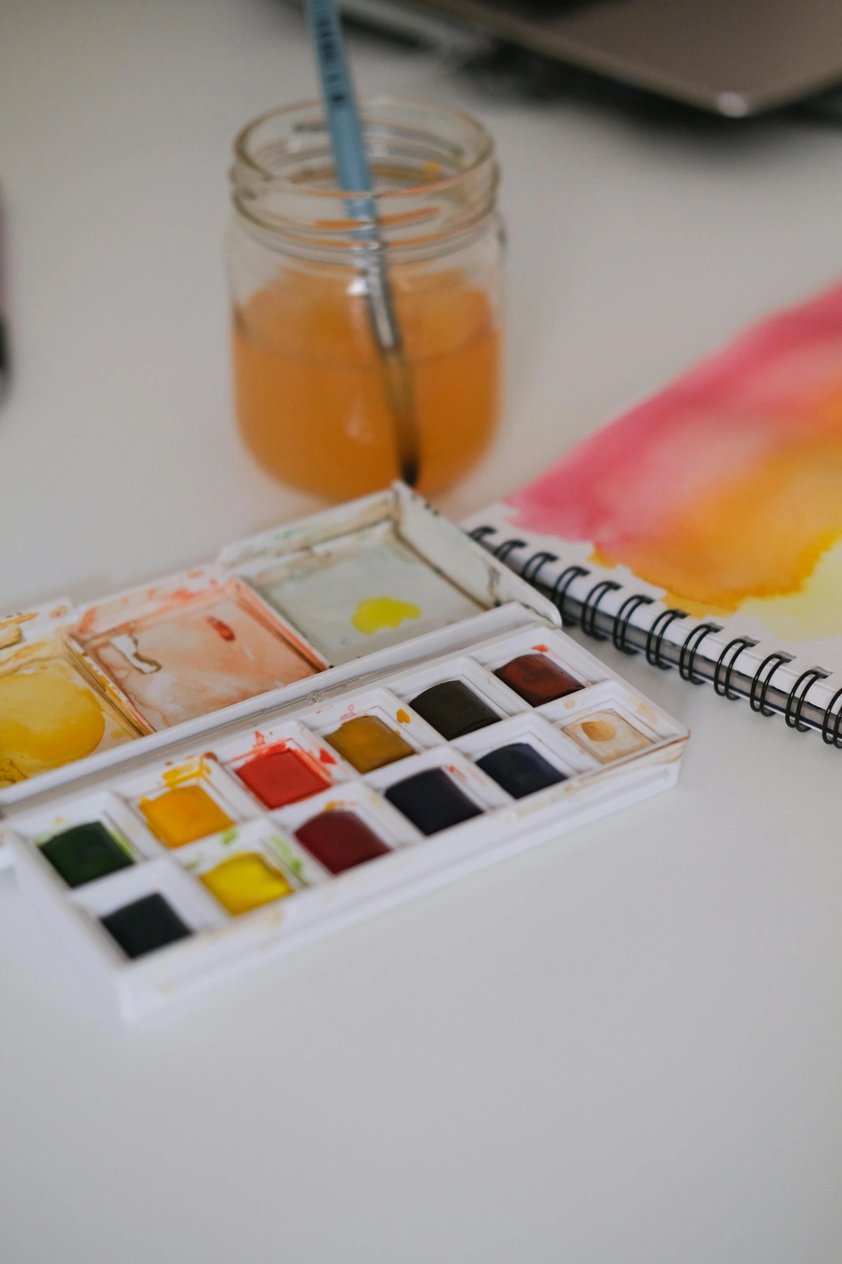 A watercolor paint set with various colors, a jar of water, and a paintbrush, alongside a sketchbook with watercolor swatches.