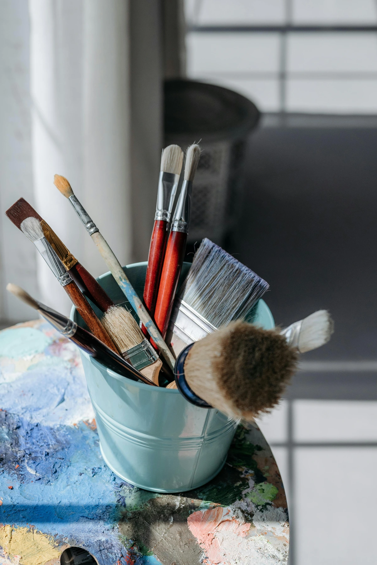 Synthetic paint brushes in a blue bucket for painting tutorials and art supplies on a paint-splattered table.