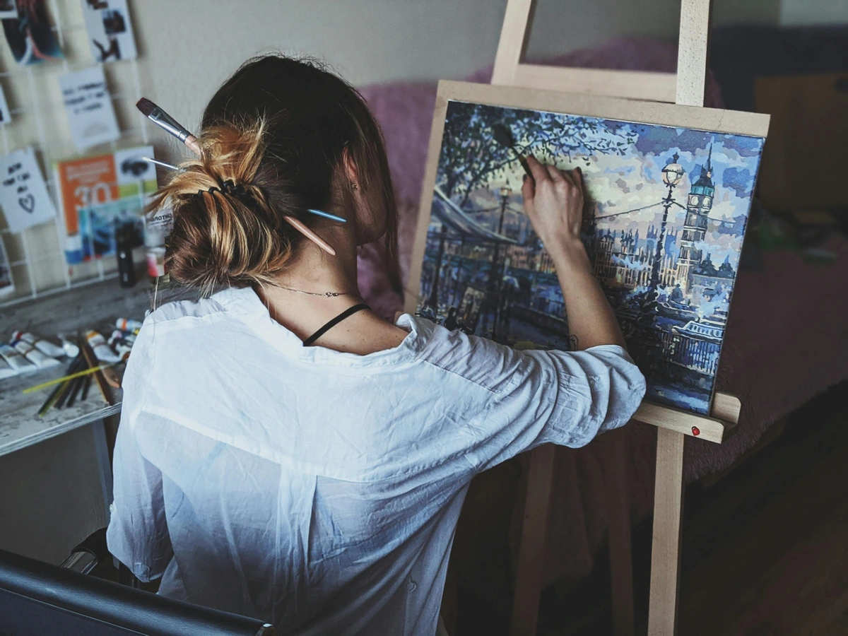 Woman Painting in Art Studio with Brushes in Hair | Cityscape Painting on Easel | Creative Process Free Stock Photo