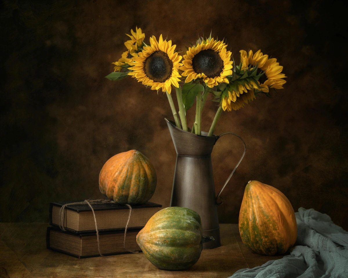 A still life arrangement featuring a bouquet of sunflowers in a metal pitcher, two old books tied with twine, and three gourds on a wooden surface against a textured brown background, emblematic of abundant fruit pieces.