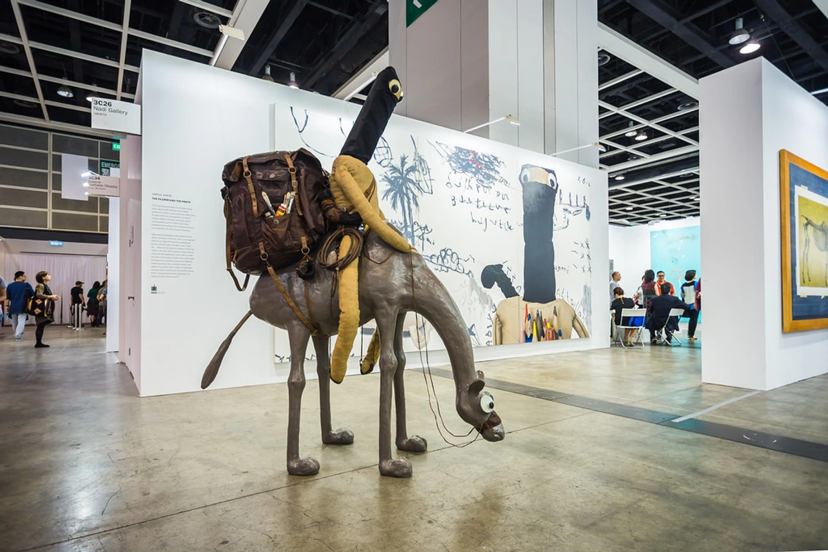 Mixed media sculpture of a camel-like creature with a backpack and a figure on its back, displayed at an art exhibition.