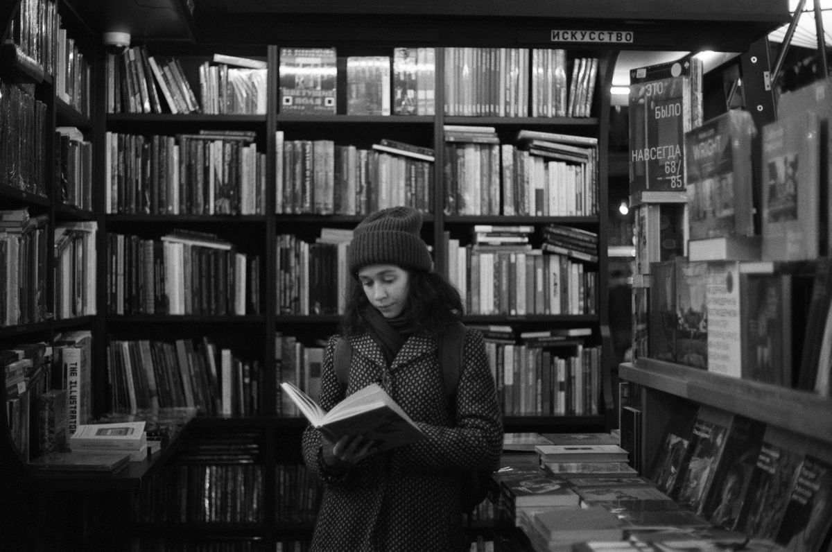 A grayscale photo of a young woman wearing a beanie and a patterned coat, engrossed in reading a book while standing in a library aisle filled with bookshelves.