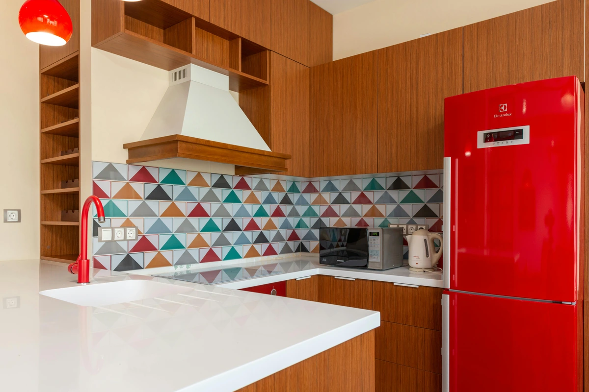Modern kitchen interior featuring a vibrant, colorful geometric tile backsplash, a sleek white countertop with a red faucet, and a bright red retro-style refrigerator.