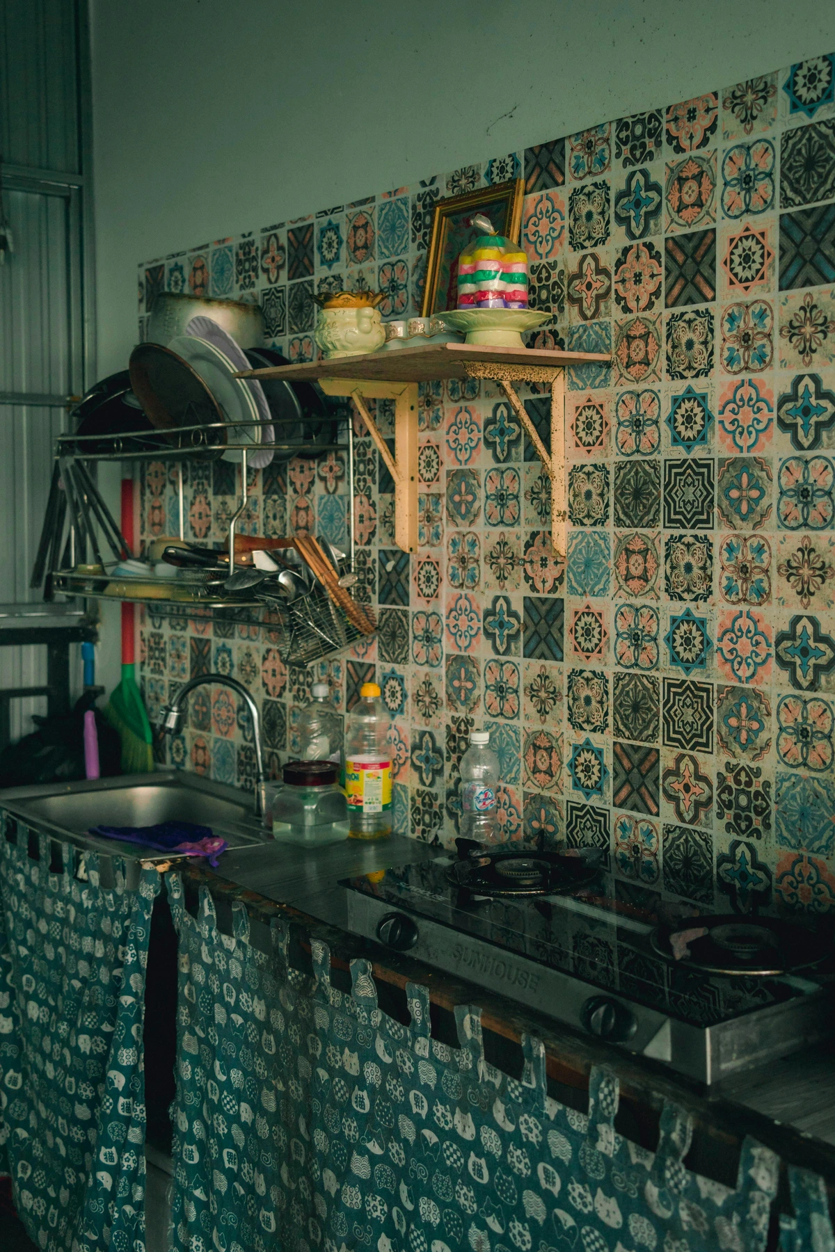 A kitchen sink and stove area with a colorful, patterned tile backsplash and a decorative curtain.
