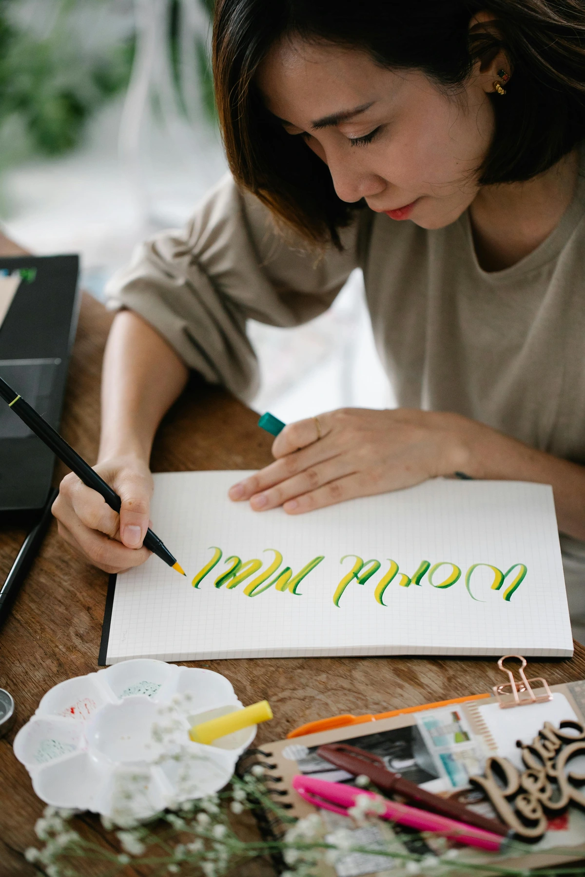 Pretty woman practicing calligraphy on a notebook with colorful pens and art supplies on a wooden table.