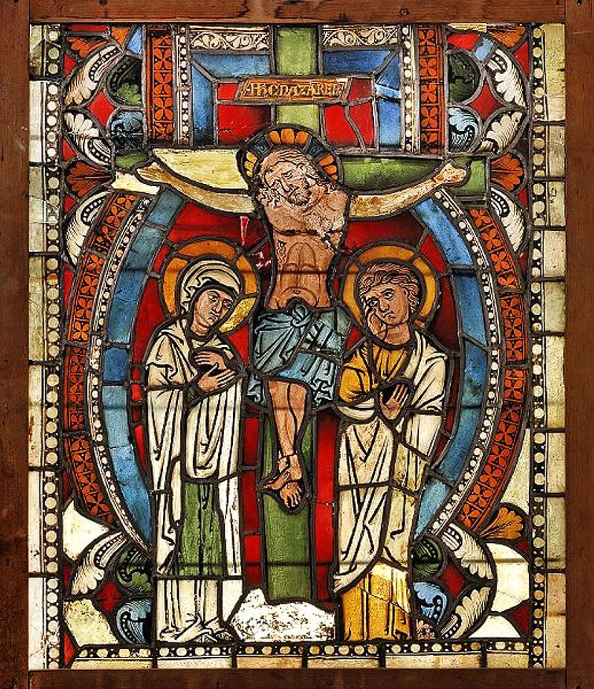 Medieval stained glass depicting the crucifixion of Jesus Christ, with Mary and John the Evangelist at the foot of the cross. The inscription above Jesus reads 'Iesus Nazarenus Rex Iudaeorum'.
