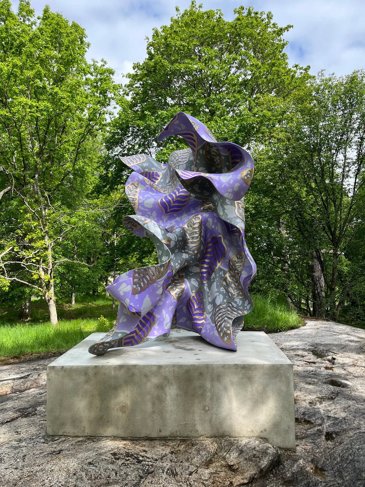 Yinka Shonibare CBE's 'Wind Sculpture in Bronze I' is a vibrant, flowing sculpture with purple and gold patterns, displayed outdoors on a concrete pedestal amidst lush greenery.