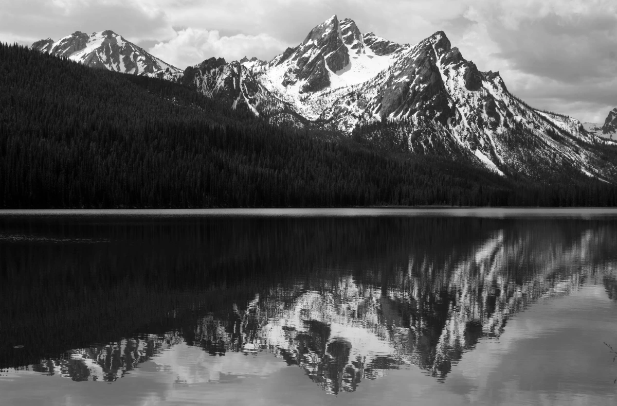 McGown Peak reflected perfectly in the calm waters of Stanley Lake, surrounded by dark evergreen forests under a dramatic cloudy sky, captured in black and white.