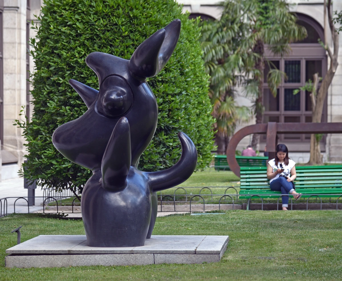 Joan Miró's 'Lunar Bird' sculpture in the courtyard of the Reina Sofía Museum, with a woman sitting on a bench in the background.