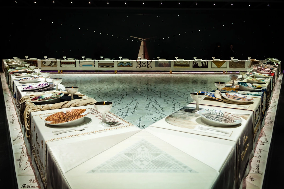 A triangular table setting for Judy Chicago's iconic feminist art installation, The Dinner Party, featuring elaborate place settings with unique plates and goblets, symbolizing historical women.
