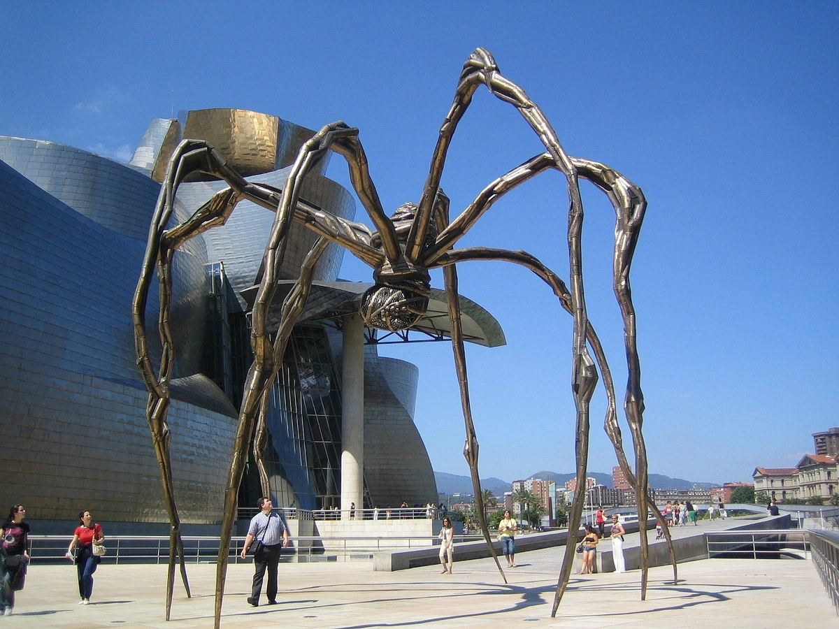 The iconic 'Maman' spider sculpture by Louise Bourgeois stands in front of the metallic, wave-like facade of the Guggenheim Museum Bilbao under a clear blue sky.