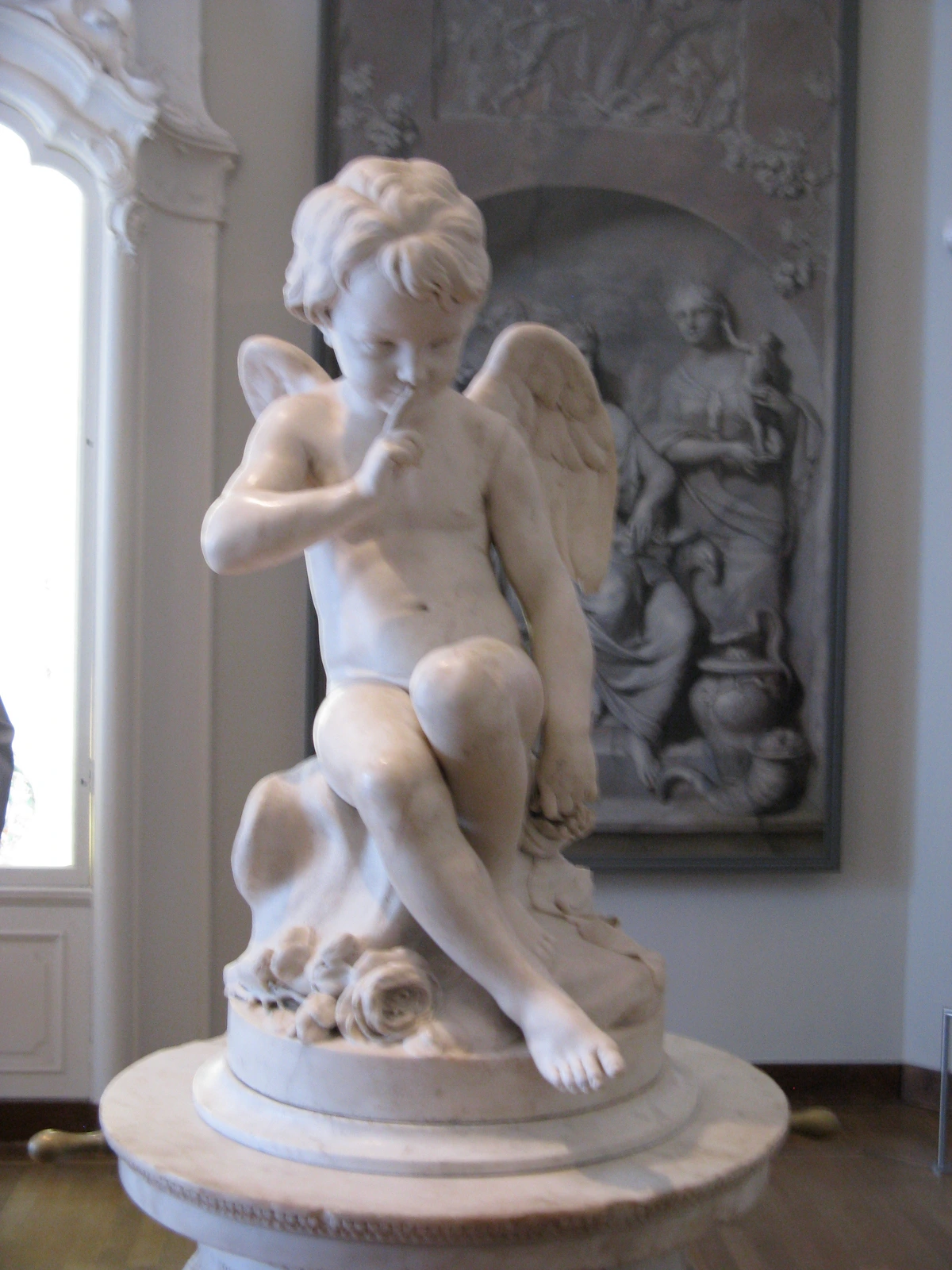 Etienne Maurice Falconet's marble sculpture of Cupid with wings, finger to lips, sitting on a pedestal adorned with roses, in the Rijksmuseum.
