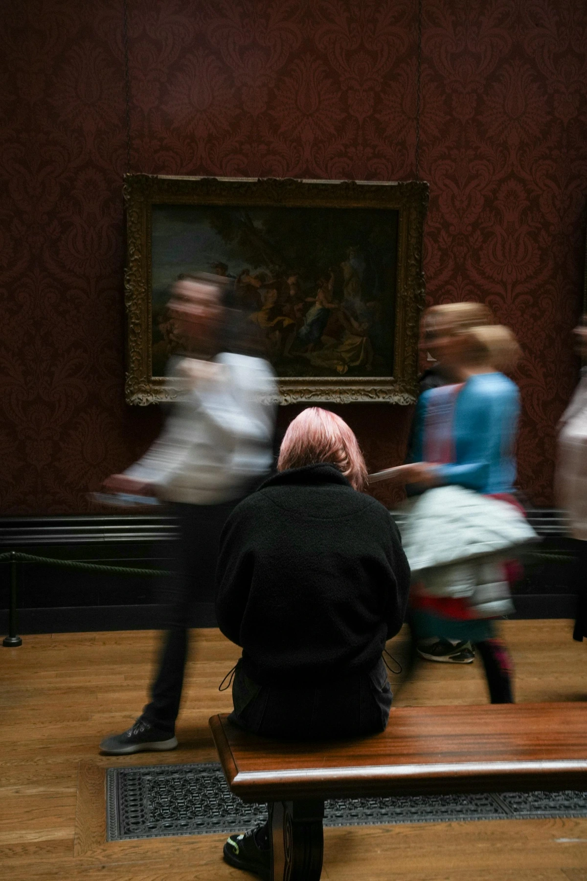 A contemplative individual examining artwork in a gallery with blurred visitors in the background. Natural midday lighting emphasizes the subject's thoughtful expression and the gallery's classical decorum, evoking a serene atmosphere of art appreciation.