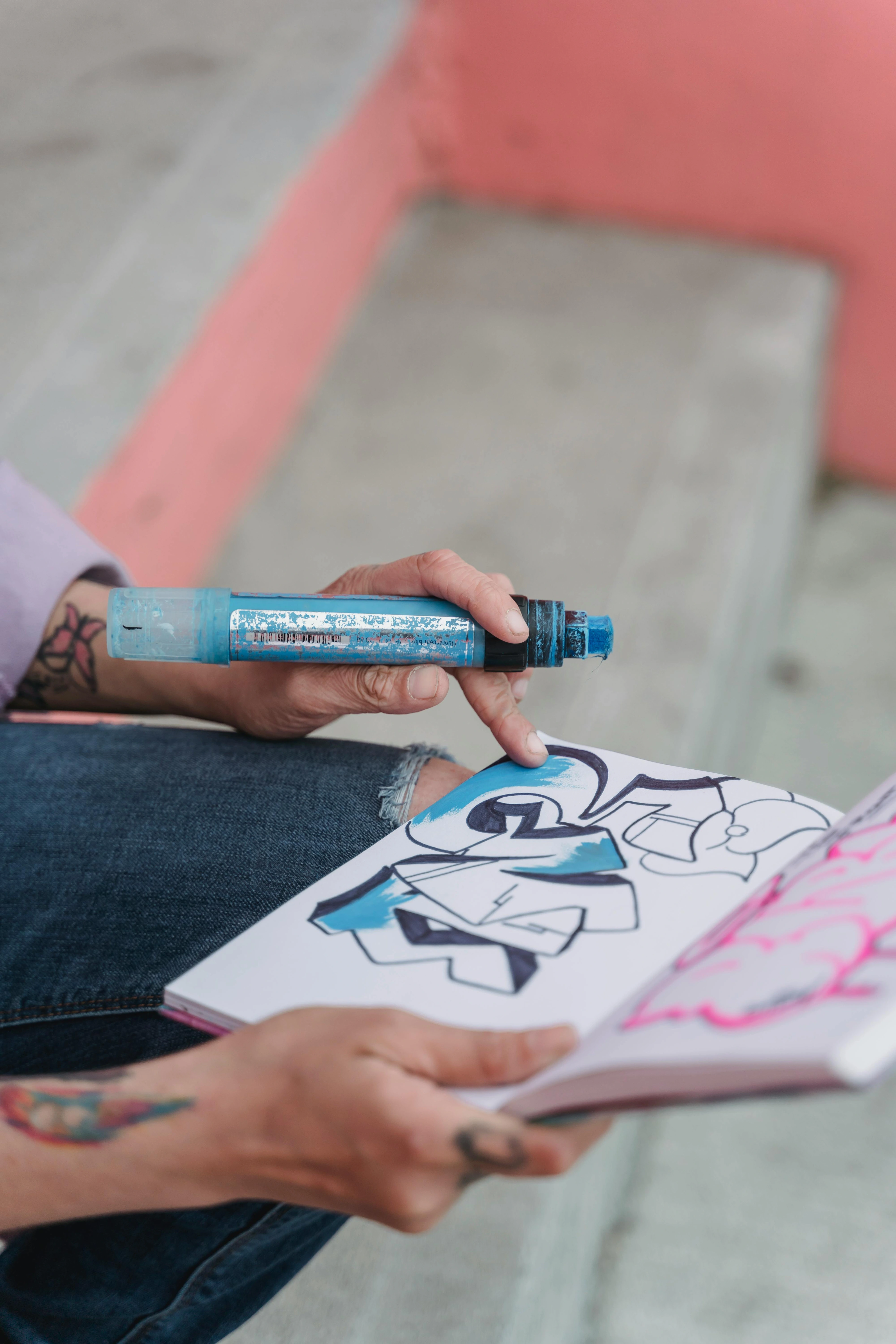 Artist's hands holding a blue Posca pen and drawing graffiti art in a sketchbook