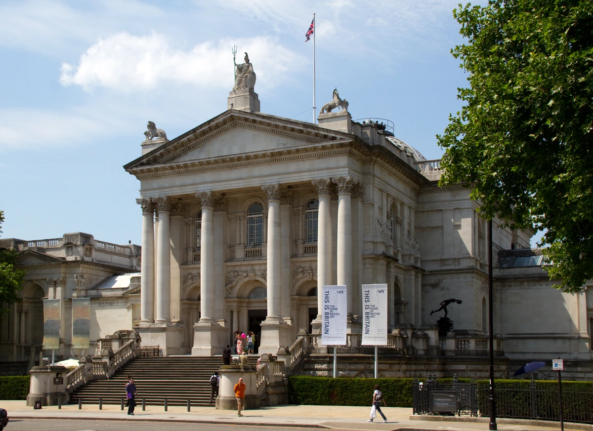 The grand neoclassical facade of Tate Britain in London, featuring a prominent portico with columns, steps leading to the entrance, and statues on the roof, under a blue sky with clouds.