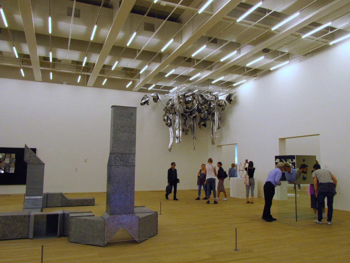 Visitors observing a large metal installation hanging from the ceiling in the Tate Modern Switch House exhibition space.